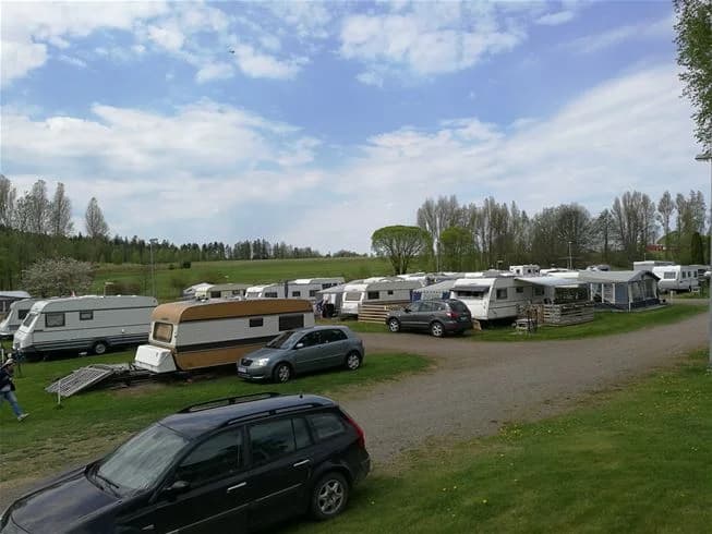 En bild över Degernäs camping som visar flera husbilar och husvagnar parkerade på en grusplan. Området är omgivet av grönska och träd, vilket skapar en avkopplande campingmiljö.