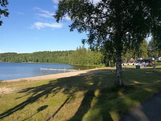 En avkopplande scen vid Degernäs Camping med en sjö i bakgrunden, omgiven av grönska. Parkbänkar och andra utomhusmöbler syns längs strandkanten, vilket skapar en inbjudande plats för besökare att njuta av naturen.