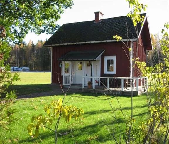 En pittoresk stuga med rött tak och ljusa väggar i en lantlig miljö på Sandaholm restaurang & camping. Rundom stugan finns gröna ytor och träd, vilket skapar en harmonisk och inbjudande atmosfär.