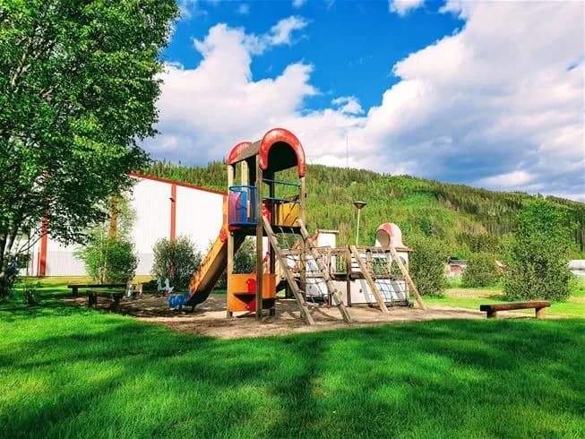 En lekplats med en rutschkana och annan utomhuslekutrustning, omgiven av grönskande gräsmatta och trädgårdsmiljö, vid Sysslebäcks stugby & fiskecamping.