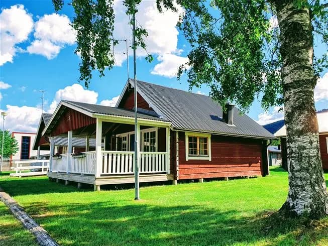 En bild av en pittoresk stuga med verandatak, del av Sysslebäcks stugby och fiskecamping, omgiven av grön gräsmatta.