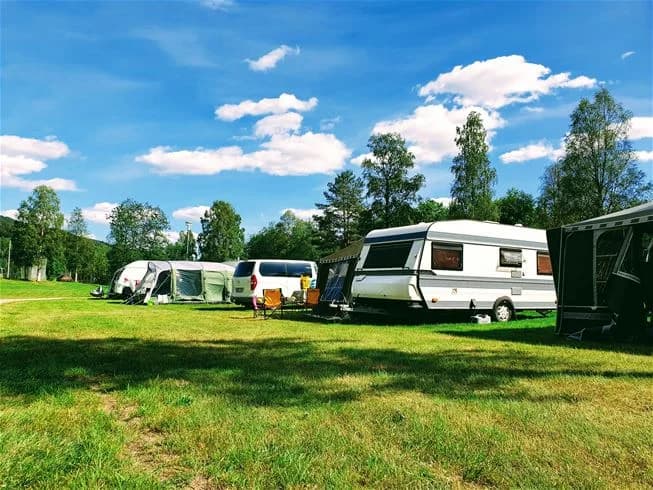 En grönskande campingplats i Sysslebäcks stugby & fiskecamping, omgiven av träd och gräs. Här syns husvagnar och semitrailer-lastbilar parkerade på den öppna ängen.