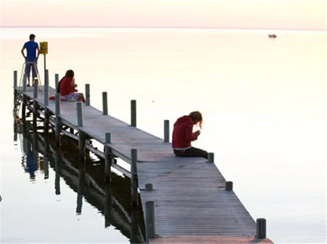 En träbrygga leder ut över havet med människor som promenerar i den natursköna omgivningen vid Lundegård Camping & Stugby, vilket ger en känsla av semester och avkoppling.