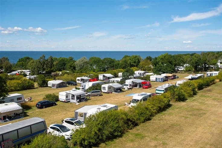 Flygbild av Lundegård Camping & Stugby med en kustlinje som sträcker sig längs ett azurblått hav. Sandstranden omges av gröna skogsområden som utgör campingen, och vågorna rullar in mot stranden. Solig och inbjudande atmosfär.