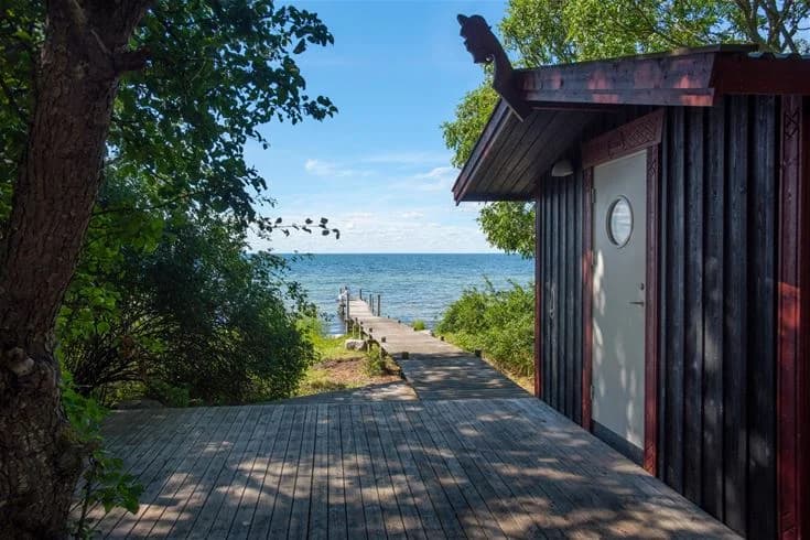 Bild av Lundegård Camping & Stugby vid kusten, där en träspång leder mot ett havslandskap. Solen skiner genom trädkronor och skapar skuggspel på marken.