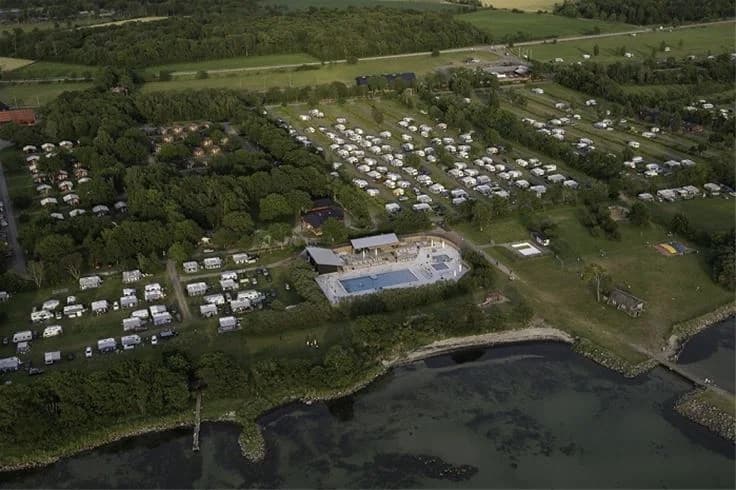 Flygfoto över Lundegård Camping & Stugby som visar en grönskande landskapsvy med hus och stugor utspridda över området. Campingplatsen är omgiven av natur med parker och små byar i bakgrunden.