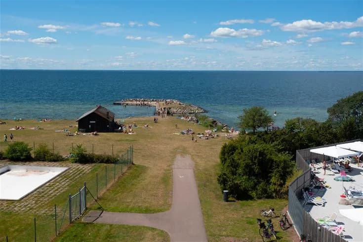 En solig dag vid kusten med utsikt över havet vid Lundegård Camping & Stugby. Sandstranden sträcker sig längs viken med lummig grönska i bakgrunden. Perfekt för en avkopplande semester vid vattnet.