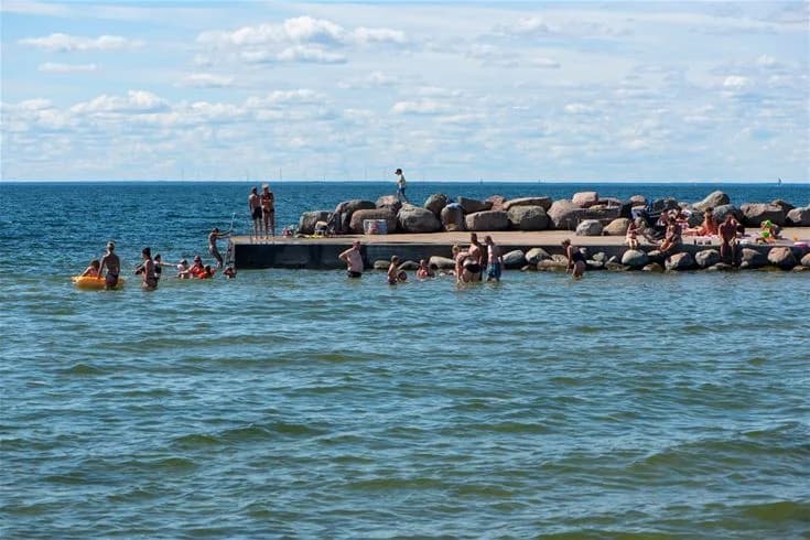 En grupp människor njuter på stranden vid Lundegård Camping & Stugby, med havet i bakgrunden och vågor som bryter lätt mot kusten. Perfekt för avkoppling och semesteraktiviteter vid havet.