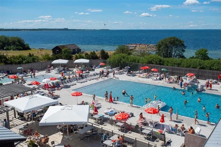 En solig dag vid Lundegård Camping & Stugby med besökare som njuter av poolområdet. Solstolar är uppställda kring poolen och människor kopplar av och semestrar i det soliga vädret.