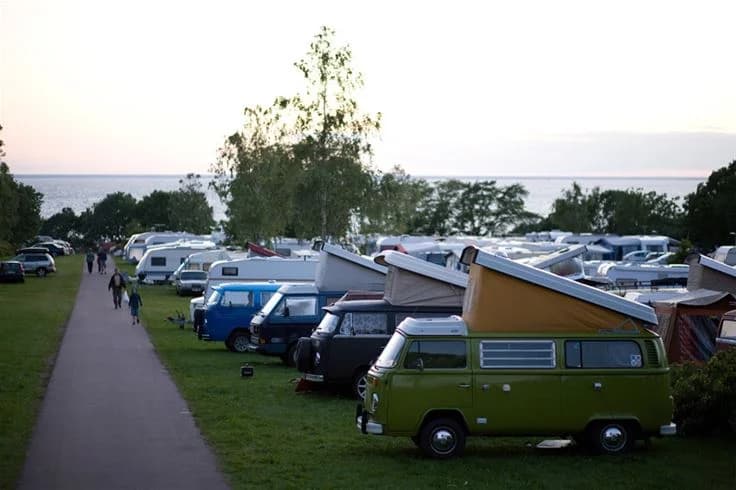 En bild av en klassisk Volkswagen husbil parkerad vid Lundegård Camping & Stugby. Husbilen står bland andra fordon på en grusad parkeringsplats, omgiven av grönska i en campingmiljö.
