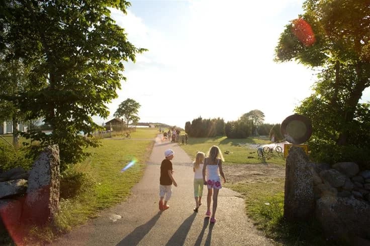 En grön gräsmatta vid Lundegård Camping & Stugby med människor som njuter av fritidsaktiviteter under en somrig dag, omgivna av molniga himlar och frodig växtlighet.