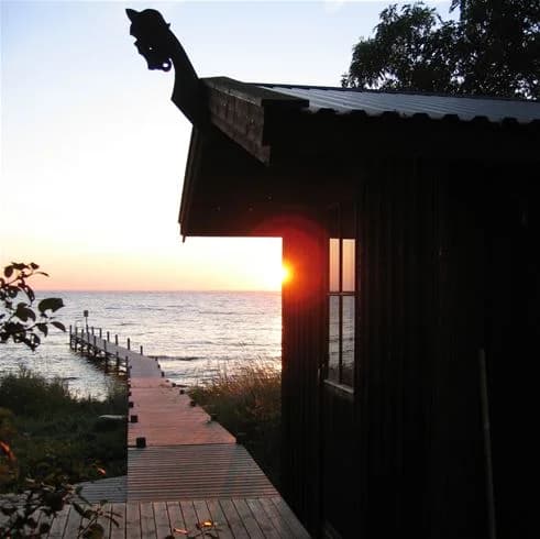 En träbrygga sträcker sig ut över det stilla havet i skymningsljus, omgiven av kustlandskap och trädlika former vid Lundegård Camping & Stugby.