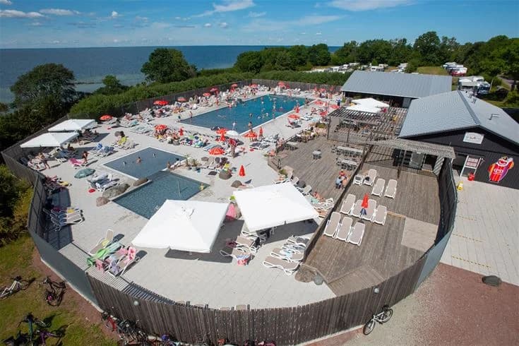 En översiktsbild av Lundegård Camping & Stugby med en stor utomhuspool omgiven av solstolar och parasoller, omgiven av grönska och campingområden för en avkopplande semesterupplevelse.