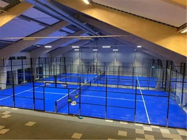 En inomhushall med hållbart golv, en stor simbassäng och ett nät för racketsport. Utrymmet verkar vara avsett för sportaktiviteter och fritid, en del av Lundegård Camping & Stugby.