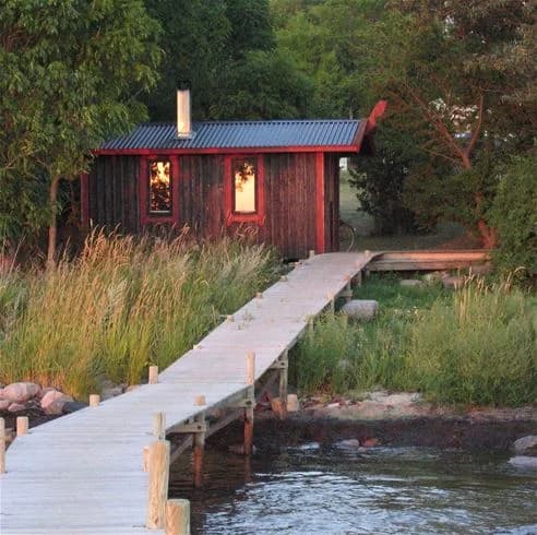 En idyllisk stuga av trä i ett lantligt område vid Lundegård Camping & Stugby, omgiven av en sjö med brygga och gångväg av plankor i förgrunden.