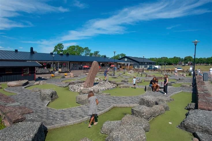 Bild av en solig parkmiljö vid Lundegård Camping & Stugby med stensatta gångvägar, skuggande tak, och områden för rekreation och vila.