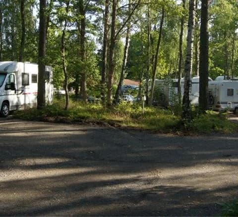 En husvagn och en husbil parkerade under trädens skugga på en grusuppfart vid Läckö Strand Camping, omgivna av grönskande skogsområde.