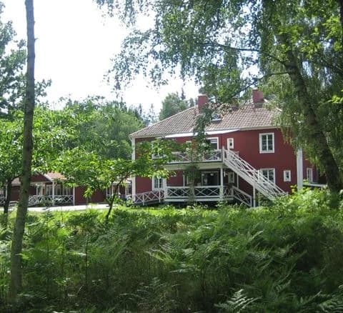 Alt-text: En lantlig campingplats vid Läckö Strand med stugor och gårdsplan, omgiven av skog och grönska.