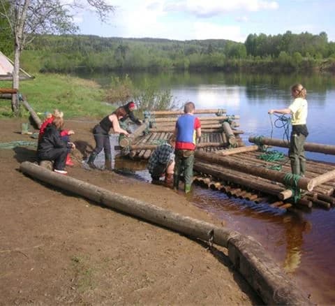 En stillsam flod sträcker sig vid Klarälvens camping, omgiven av frodig grönska. Besökare syns njuta av naturen och olika fritidsaktiviteter vid vattnet.