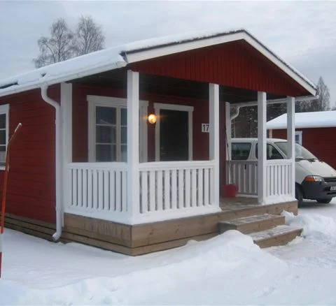 En trästuga med veranda på Klarälvens camping, omgiven av grönska. Stugan har träpaneler och en utomhusstruktur som ger en rustik känsla.