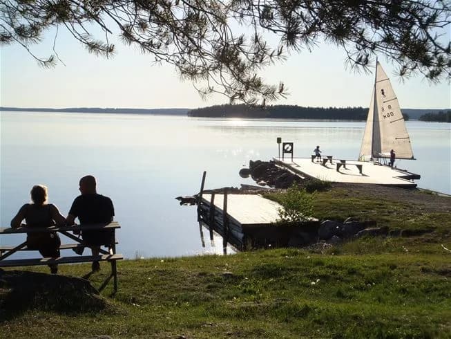 En fridfull sjö vid en campingplats i Tiveden, med båtar förtöjda vid en brygga och omgivande natursköna stränder.