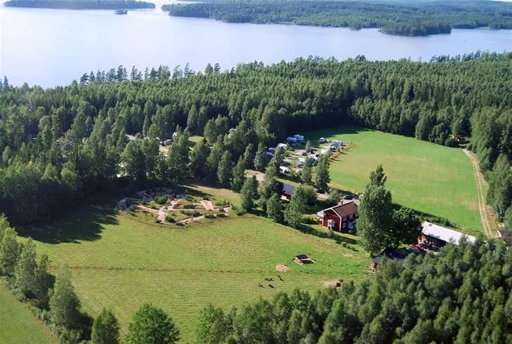 Flygbild över en frodig campingplats i Tiveden där träd och gröna växter omfamnar små stugor och gräsmattor i en lantlig miljö.