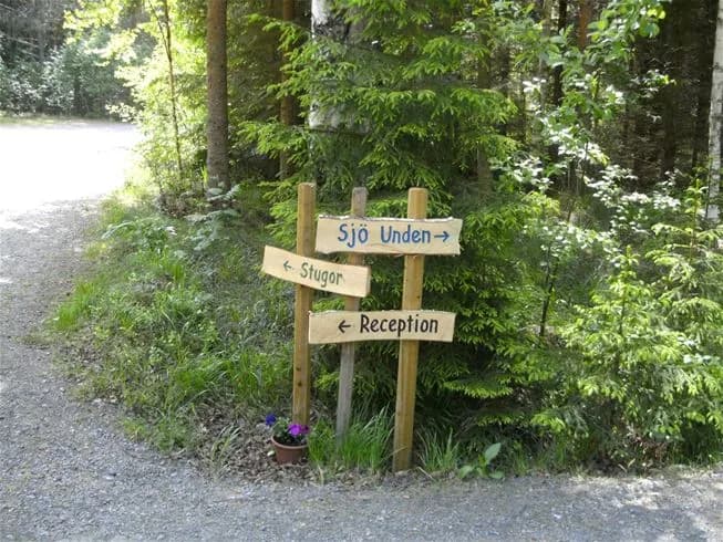 Bild på en camping vid Tiveden, omgiven av grönskande skog. En träskylt står bland buskar och markväxter, och indikerar närheten till ett naturreservat.