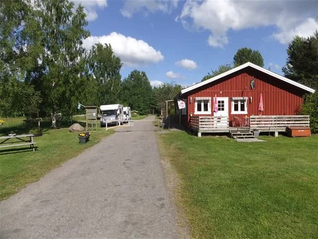 En grön och vidsträckt äng med en stuga i bakgrunden omgiven av parkbänkar, belägen vid camping Tiveden.