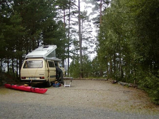 En kompakt husbil är parkerad på en campingplats omgiven av skog, med en takmonterad lastbärare och öppna bildörrar som skymtas.