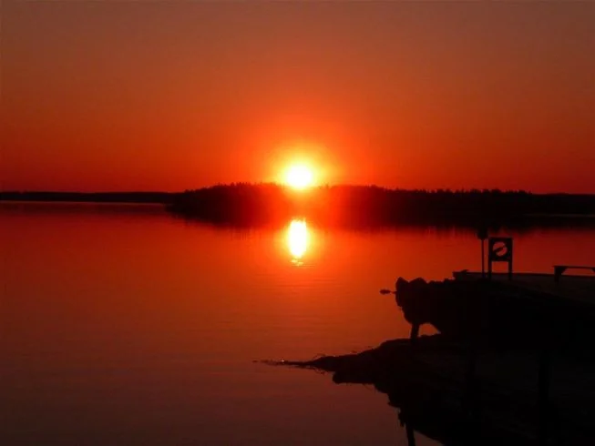 En solnedgång över en vattenresurs i Tiveden, med horisonten i bakgrunden. Himlen är färgad i orange och rosa nyanser som speglas i det stilla vattnet, vilket skapar en lugn och naturskön atmosfär.