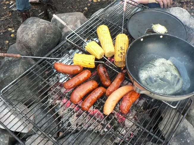 En grill utomhus med korvar som grillas över öppen eld, omgiven av gröna omgivningar inom campingområdet Tiveden.