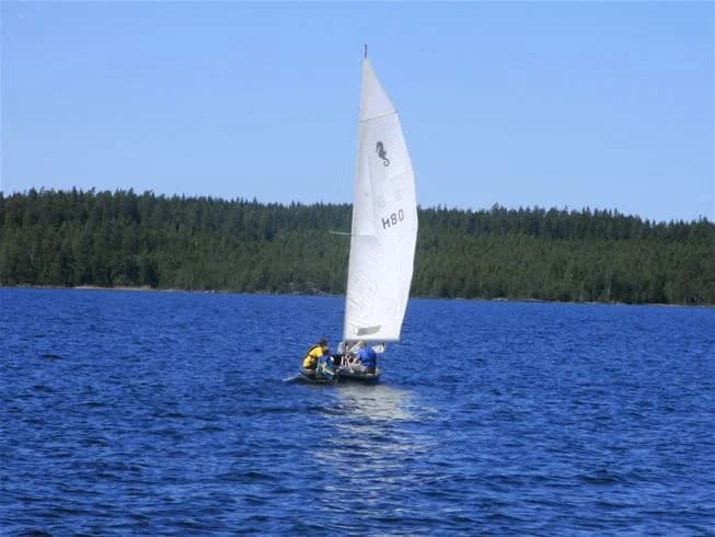 En segelbåt med hissade segel glider fram över en lugn vattenyta, omgiven av skog och natursköna vyer i Tiveden.