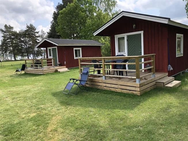 Bild av Malexander Camping som visar en grönskande campingplats med en logstuga bland gräsytor och buskar. I bakgrunden syns träd och en mobil hemvagn. Avkopplande och naturnära miljö för besökare.