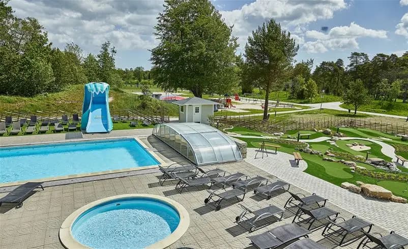 En solig dag vid Sudersand Resort: Besökare njuter av en stor swimmingpool omgiven av välskötta gräsmattor och frodig trädgårdslandskap, vilket skapar en avkopplande miljö för rekreation och fritid.