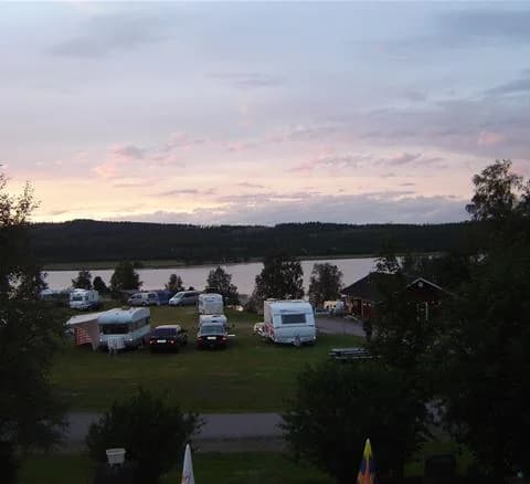 En campingplats vid en pittoresk sjö med en husbil och en smal bil parkerade i förgrunden, omgiven av naturskön miljö och kuperat landskap i bakgrunden.