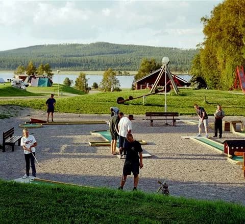 En grupp barn som leker på en grön gräsmatta vid Särna camping, omgivna av träd och picknickbord, en solig dag för utomhusaktiviteter och avkoppling.