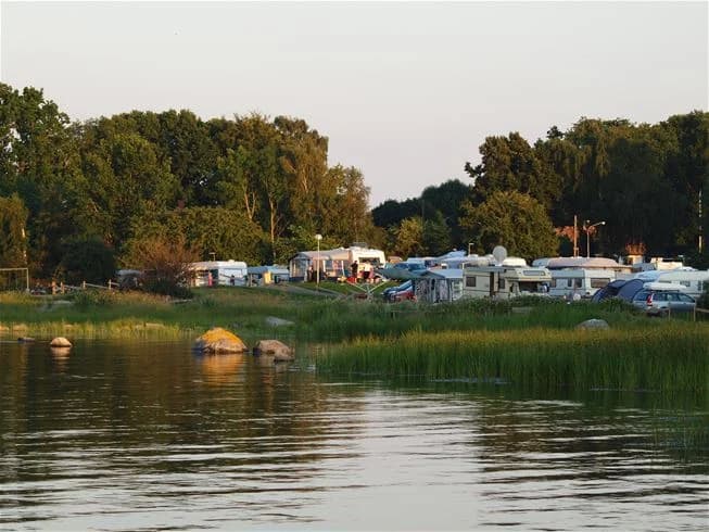 En somrig campingplats med tält och husvagnar vid en sjö, med strand och grönskande omgivningar.