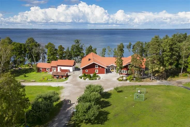 Alt-text: Bild av en lantlig stuga omgiven av grönska vid Hjälmargården & Läppe Camping, med ett pittoreskt bostadsområde i bakgrunden.