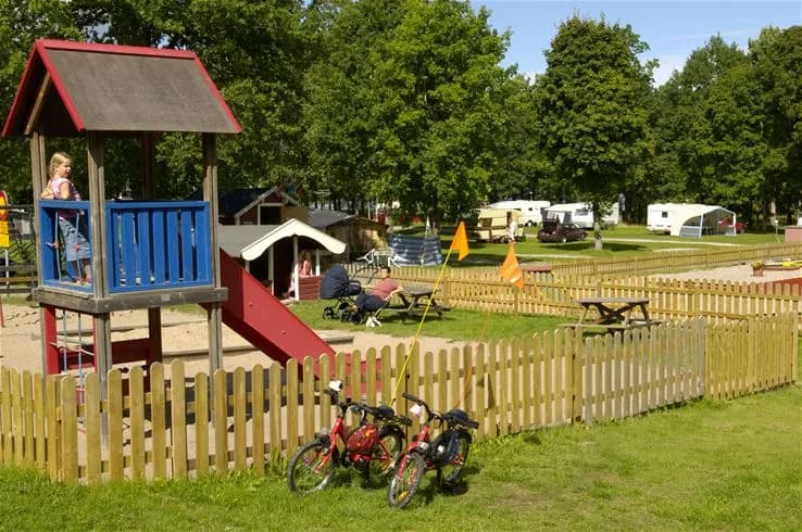En grönskande utomhusmiljö på Hjälmargården & Läppe Camping med lekutrustning för barn, tält och skuggiga områden.