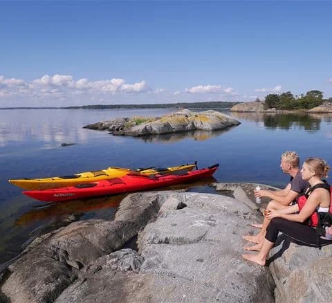 En grupp människor paddlar i kajaker på Gålö Havsbad, omgivna av kustlinje och vacker havsmiljö. Flera båtar syns i bakgrunden under en klar himmel.