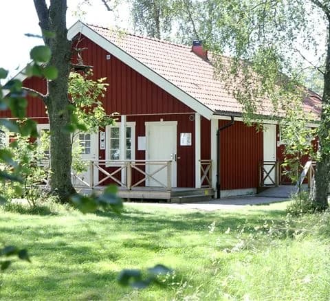 Ett rött stugliknande hus vid Gålö havsbad, omgiven av grönska med en välskött gräsmatta och en veranda.