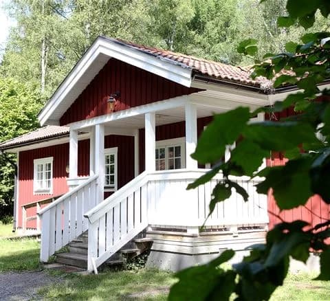 En mysig stuga med träfasad och veranda, beläget vid Gålö havsbad, omgiven av grönska.