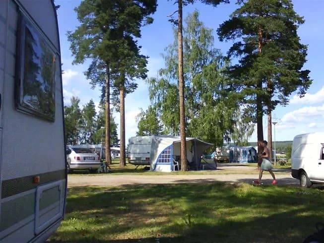 En campingplats med parkerade husbilar och husvagnar under skuggande träd, omgivna av öppna gräsytor och parkeringsplatser.
