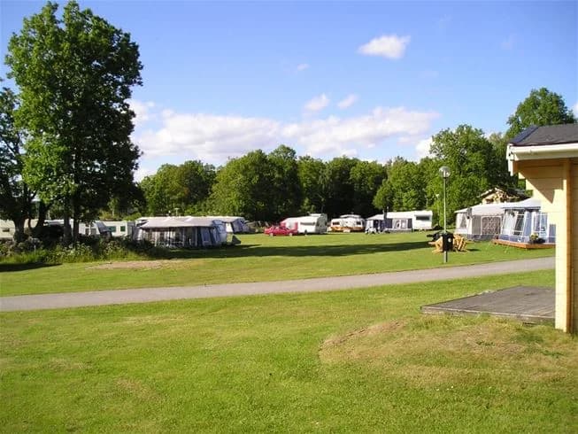 En campingplats med parkerade husbilar och bilar på en grön gräsmatta, omgiven av träd och ett område med skugga. Himlen är lätt molnig och platsen har en fridfull, naturnära atmosfär.