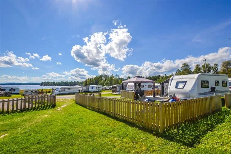 Panoramabild av Charlottenberg camping & stugor - Haganäset, med en vacker utsikt över ett reservoar och omgivande grönska. Flera husvagnar och campingfordon syns parkerade, omgivna av ett kuperat landskap med cumulusmoln på himlen.