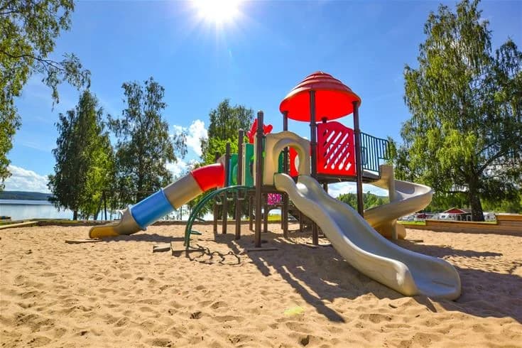 En somrig campingplats med utelekplats, pool och trädgårdsmöbler i skuggan, omgiven av grönska.