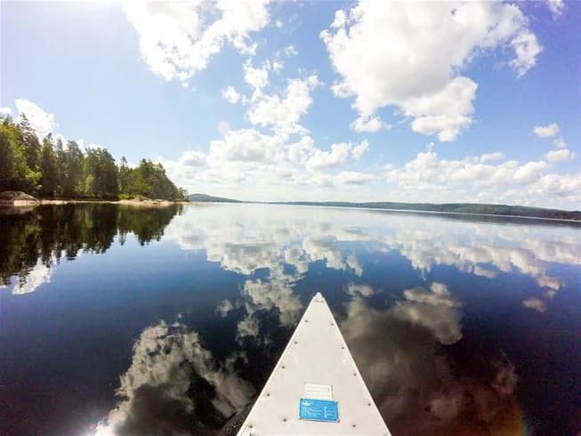 En klarblå himmel speglas i en lugn sjö vid Charlottenberg camping & stugor - Haganäset. Några moln syns vid horisonten och en båt kan skymtas på vattnet.