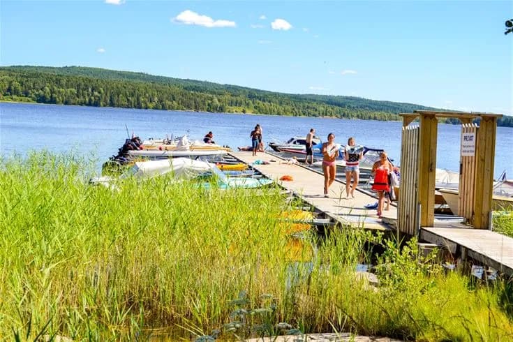 En somrig bild av Charlottenberg camping & stugor - Haganäset med en sjö i förgrunden där en båt ligger förtöjd vid en brygga. I bakgrunden syns stranden och natursköna omgivningar som inbjuder till rekreation.