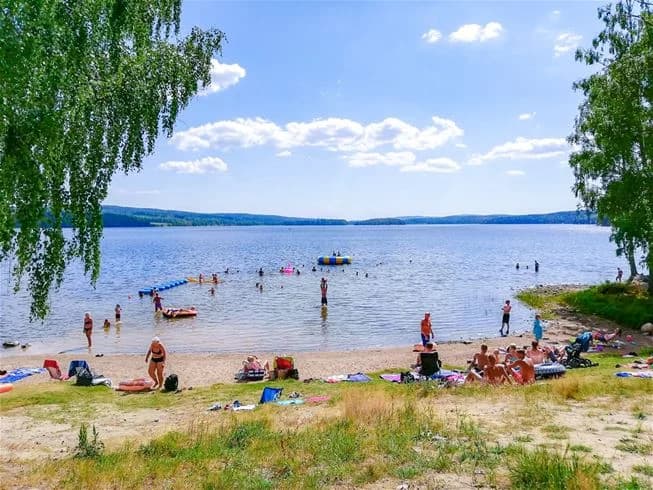 En somrig bild över Charlottenberg camping & stugor - Haganäset, med vatten i förgrunden och sandstrand i bakgrunden. Människor njuter av sol och bad längs kusten.