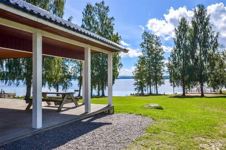 Bild på Charlottenberg camping & stugor - Haganäset med vidsträckt grön gräsmatta, träd som ger skugga, och en paviljong bland trädgårdsbyggnader. Området är parkliknande med närhet till vatten, vilket ger en fridfull atmosfär i naturskön miljö.